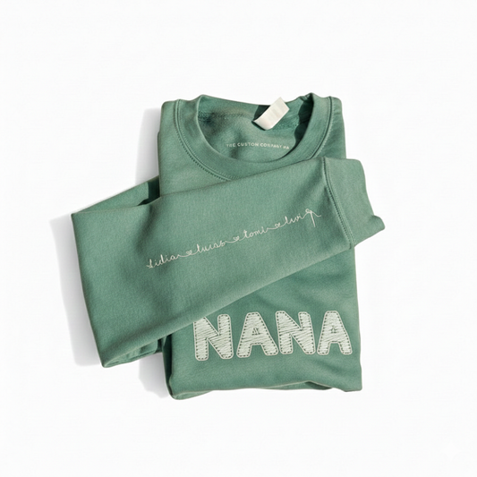 A high-quality photo of a bespoke sage-colored sweatshirt featuring the word "Nana" in a tactile, 3D cross-stitch effect. The design is centered on the chest, showing a warm and modern aesthetic.