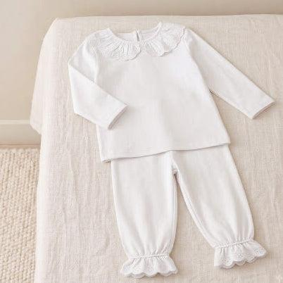 A white two-piece baby outfit consisting of a long-sleeve top with a ruffled Peter Pan collar and matching pants with ruffled cuffs, laid flat on a beige fabric-covered surface.