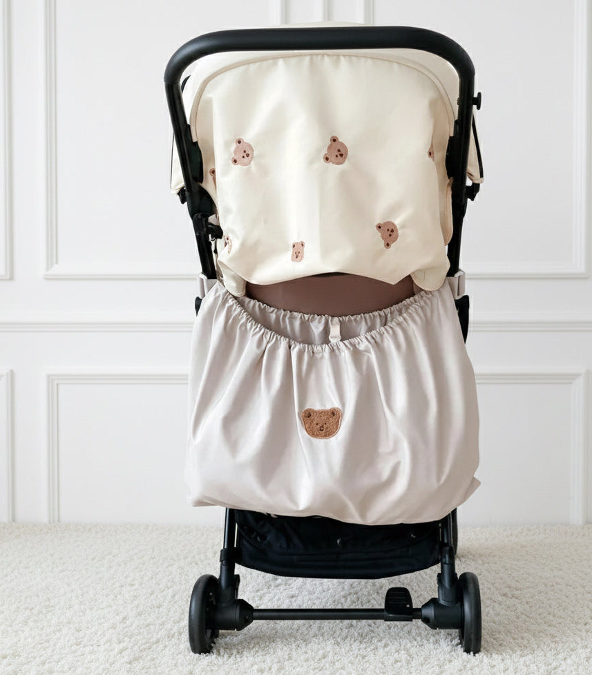 Neutral personalised stroller bag with embroidered teddy bear design attached to a black pram, featuring a large elasticated storage pocket and soft cotton finish in a minimalist white room.