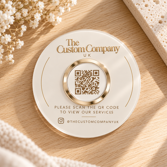 A round white and gold branded acrylic disc from The Custom Company UK, featuring a central QR code and text inviting users to scan the QR code to view their services. The card is styled on a light wood surface.