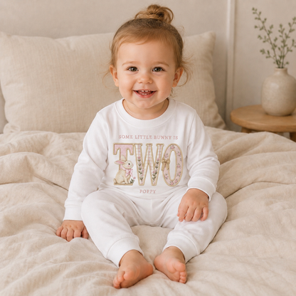 Young girl wearing personalised white pyjamas featuring embroidered style bunny birthday design reading Some Little Bunny Is Two