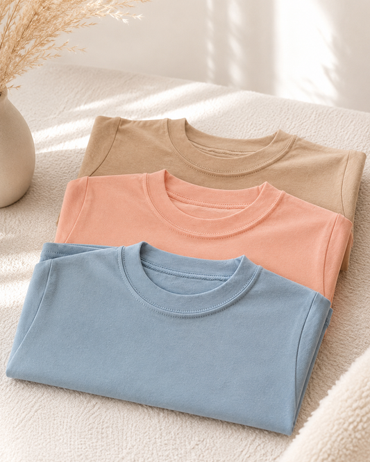 Three folded short-sleeve crewneck T-shirts in light blue, peach pink, and beige, arranged on a textured cream fabric surface with soft natural shadows and a vase of dried grass in the corner.